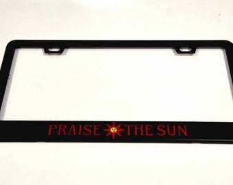 Puede incluir: Marco de matrícula negro con las palabras "PRAISE THE SUN" en letras rojas. El símbolo del sol también es rojo y amarillo. El marco tiene forma rectangular con dos orificios de montaje.