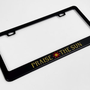 Könnte beinhalten: Schwarzer Kennzeichenrahmen mit dem Text "PRAISE THE SUN" in goldener Schrift und einer rot-gelben Sonnengrafik. Der Rahmen ist rechteckig mit einem schwarzen Rand und Befestigungslöchern.