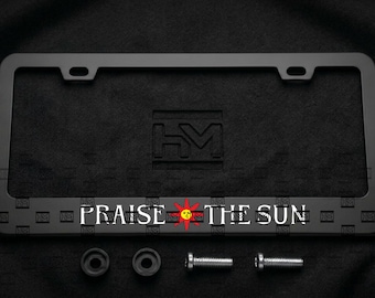 Puede incluir: Un marco de matrícula rectangular de color negro mate con el texto "PRAISE THE SUN" en letras blancas y un pequeño icono de sol rojo y amarillo en el centro. El marco incluye dos arandelas negras y dos tornillos de metal plateado.