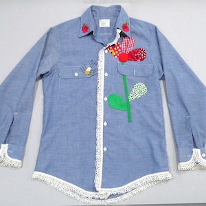 May include: Light blue button-down shirt with long sleeves, adorned with a large flower applique and an insect. The shirt features white lace trim along the placket, cuffs, and hem, and embroidered red roses on the collar.