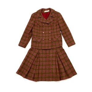 May include: A vintage two-piece suit in a maroon and olive green plaid pattern. The jacket is double-breasted with four buttons and two front pockets. The skirt is pleated. The suit is displayed on a white background.