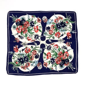 May include: Square navy blue scarf with a white floral design. The scarf features a repeating pattern of flowers in shades of red, blue, and green. The scarf has a white border.