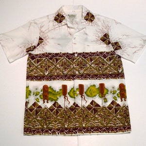 May include: White short-sleeve shirt with a colorful pattern. The shirt features a geometric design in shades of brown and green, with additional brown and green tiki-like figures and tree branch patterns. The shirt has a collared neckline and button-up front.