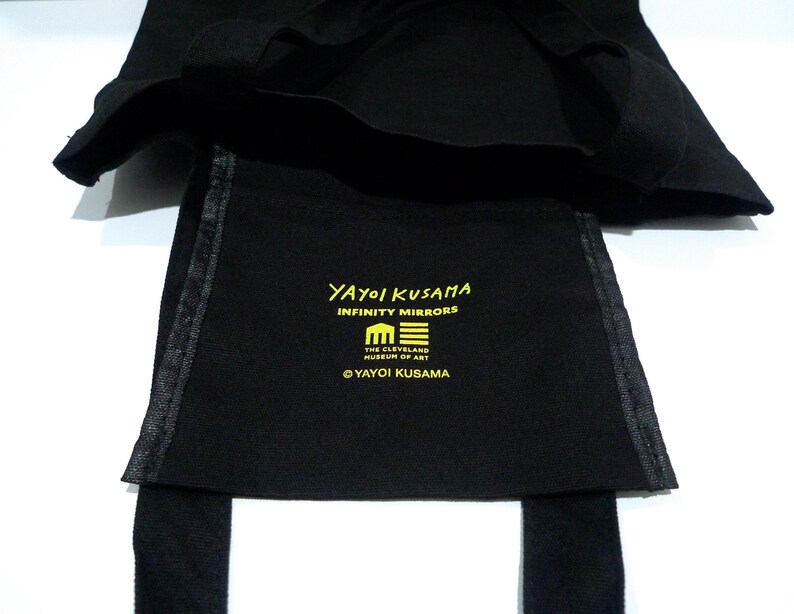 May include: Black tote bag with the text "YAYOI KUSAMA INFINITY MIRRORS" in yellow. The bag also features the emblem of The Cleveland Museum of Art in Ohio and a pumpkin graphic. The bag has straps.