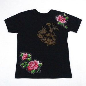 May include: Black t-shirt with floral and bird designs. The shirt features a large gold-colored magnolia and bird graphic, along with two patches of pink roses with green leaves. The text "Remember" is above the bird.