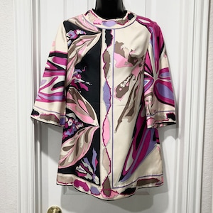 May include: A colorful tunic top with a high neckline and three-quarter sleeves. The top features a bold, abstract pattern in black, cream, pink, purple, and brown. The design is symmetrical, with a central black panel and floral-like shapes.