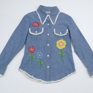 May include: A light blue denim shirt with long sleeves, a white lace collar, and white lace trim. The shirt features embroidered flower and snail appliqués in red, yellow, and blue. The shirt has a button-down front and two chest pockets.