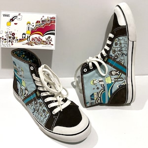 May include: Pair of high-top Vans sneakers with a unique illustrated design. The shoes feature a light blue and brown color scheme with white laces and soles. The illustration includes cartoon figures and buildings. A small card with the Vans logo is also present.
