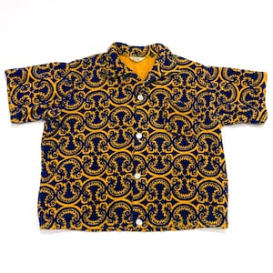 May include: A short-sleeved button-down shirt with a repeating navy blue and gold pattern. The shirt has a collar and white buttons. The interior collar is a solid gold color.