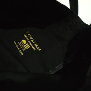 May include: Black tote bag with yellow text that reads "Yayoi Kusama Infinity Mirrors" and "The Cleveland Museum of Art". The bag has black handles and is made of a soft material with a pumpkin graphic.