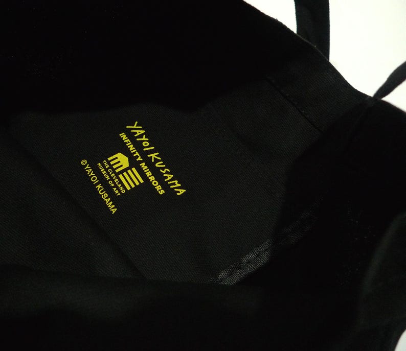 May include: Black tote bag with yellow text that reads "Yayoi Kusama Infinity Mirrors" and "The Cleveland Museum of Art". The bag has black handles and is made of a soft material with a pumpkin graphic.