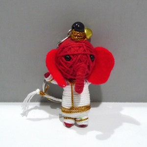 May include: A handmade red elephant keychain with large red ears, a white outfit, and gold accents. The elephant's head is made of red yarn, and it has black eyes. The keychain includes a small black hat and a gold-colored bell.