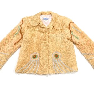 May include: A light tan jacket with a fuzzy texture and decorative embroidery. The jacket features a rounded collar, button closure, and embroidered details in peach, green, and gray. The sleeves have a unique design.
