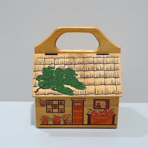 May include: Wooden house-shaped box with a handle. The roof is textured with a green tree painted on it. The front features a door, window, and flower boxes. The box is painted with orange, red, and green accents.