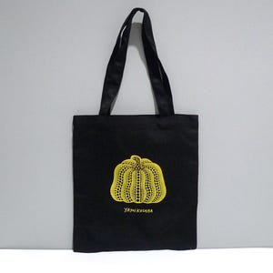 May include: Black Yayoi Kusama Pumpkin Print, Cleveland Museum of Art tote bag with a yellow pumpkin design featuring a dotted pattern. The bag has two black handles and the text "YAYOI KUSAMA" in yellow. The bag is suitable for carrying various items.