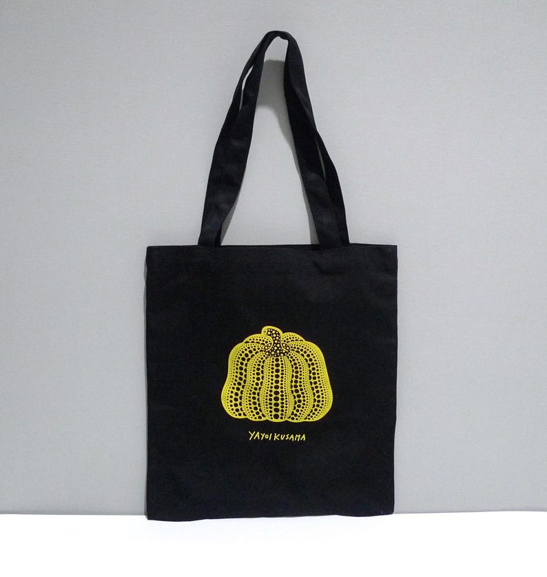 May include: Black Yayoi Kusama Pumpkin Print, Cleveland Museum of Art tote bag with a yellow pumpkin design featuring a dotted pattern. The bag has two black handles and the text "YAYOI KUSAMA" in yellow. The bag is suitable for carrying various items.