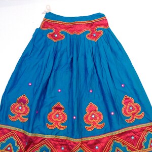 May include: A vibrant blue skirt with ornate red and gold embroidered details. The skirt features decorative mirrored accents and a wide red waistband. The design includes repeating heart-shaped patterns and geometric borders, creating a festive look.