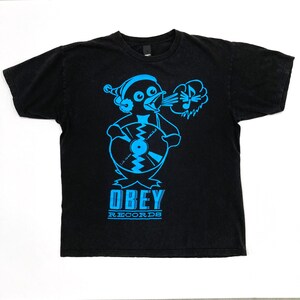May include: Black t-shirt featuring a blue graphic of a penguin wearing headphones and holding a record. The penguin is blowing musical notes. The text "OBEY RECORDS" is printed below the penguin.