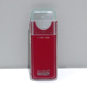 May include: A red Swatch Irony watch case with a clear plastic cover. The case is rectangular with a silver trim and the words "SWISS MADE" are printed on the front. The Swatch Irony logo is at the bottom.
