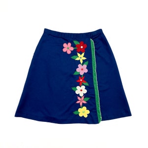 May include: A navy blue wrap skirt with a colorful floral design. The skirt features a row of appliqued flowers in pink, red, yellow, and white, with green leaves. A green trim accents the edge of the skirt.