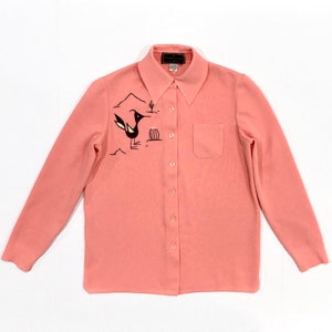 May include: A light coral-colored button-down shirt with long sleeves. The shirt features a black and white embroidered roadrunner design with desert landscape elements. A small pocket is on the left chest. The label reads "Country Casuals".