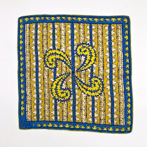 May include: Square silk scarf with a vibrant design. The scarf features a yellow and white striped background with a large, symmetrical yellow and blue floral design in the center. A blue border with a repeating pattern frames the scarf.