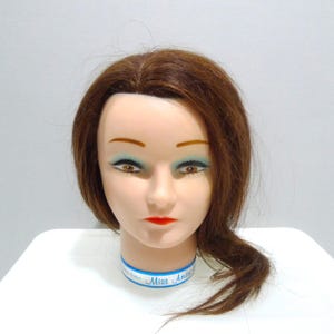 May include: A mannequin head with brown hair, blue eye shadow, and red lipstick. The mannequin has a blue and white collar that reads "Miss America". The head is on a white surface.
