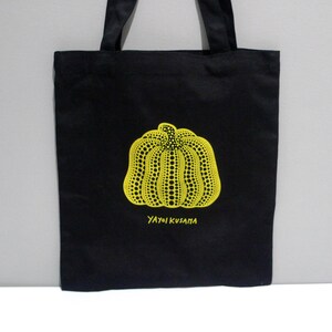 May include: Yayoi Kusama Cleveland Museum Black canvas tote bag featuring a yellow dotted pumpkin design. The text "YAYOI KUSAMA" is printed below the pumpkin. The bag has black handles and is made of a sturdy fabric.