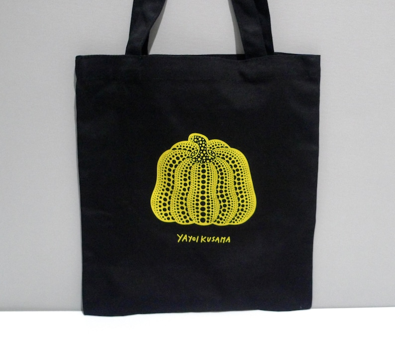 May include: Yayoi Kusama Cleveland Museum Black canvas tote bag featuring a yellow dotted pumpkin design. The text "YAYOI KUSAMA" is printed below the pumpkin. The bag has black handles and is made of a sturdy fabric.