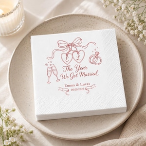 May include: A square white paper napkin featuring a red printed design of two hearts with the letters E and L, a bow, two champagne flutes, and two rings. The text reads The Year We Got Married, Emma & Lucas, and the date 05.09.2026.