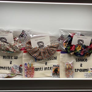 May include: Three gift sets in clear plastic packaging, each containing a scrunchie, a matching hair tie, and a canvas bag. The bags have text and stickers. Scrunchies feature floral, plaid, and patterned designs. The bags have text that reads "make up & melt", "Pinocchi & anti intel", and "tonico & traumi".