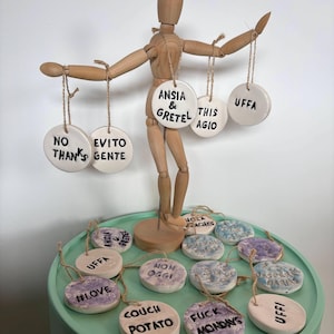 May include: A wooden artist's mannequin displays ceramic ornaments with black ink phrases. The round ornaments are strung with twine. Phrases include "No Thanks," "Evito Gente," and "Anisa & Gretel." Other ornaments have purple and blue designs.