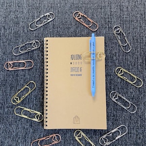 Puede incluir: Un cuaderno en espiral marrón con el texto "ADULTING DIFFICULT AF" y un bolígrafo azul. Varios clips de papel metálicos en plata, oro y oro rosa rodean el cuaderno. El bolígrafo dice "I need a six month vacation twice a year."
