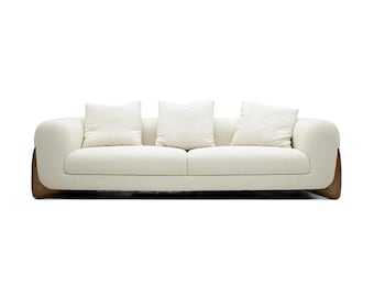 May include: A modern, cream-colored sofa with three white square pillows. The sofa has rounded arms and a wooden base. The design is minimalist and contemporary, suitable for a living room or lounge area.