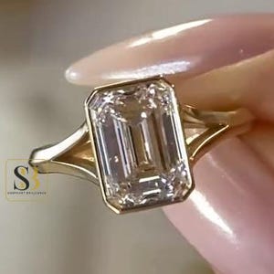 Puede incluir: Un anillo de oro con una gran piedra preciosa transparente de corte esmeralda rectangular en un engaste de bisel. La banda tiene un diseño de vástago dividido que se estrecha hacia la piedra central. Un pequeño logotipo cuadrado con SB es visible.