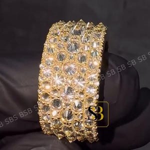 May include: A wide, gold-toned ring encrusted with numerous clear, sparkling gemstones. The ring has a thick band and a textured surface, reflecting light. The ring is held against a dark background.