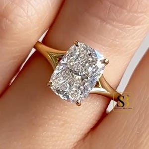 Op de afbeelding: Een gouden ring met een grote, rechthoekige, kussengeslepen heldere edelsteen die door vier pootjes wordt vastgehouden. De ringband heeft een gespleten ontwerp dat taps toeloopt naar de middelste steen. Een SB-logo is zichtbaar.