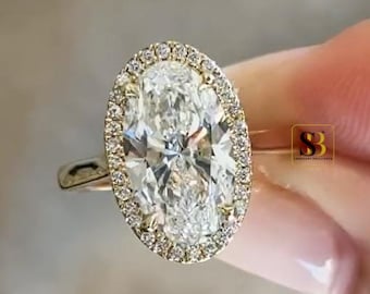 May include: A gold ring featuring a large oval-cut center stone surrounded by a halo of smaller round-cut stones. The band is thin and metallic gold. A small square logo with the letters SB is visible on the right side of the ring setting.