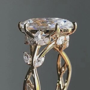 May include: A gold ring featuring a large oval-cut clear center stone held by prongs. The band is designed with intertwining metal vines and small marquise-cut clear stones set along the sides, creating a detailed, branch-like appearance.