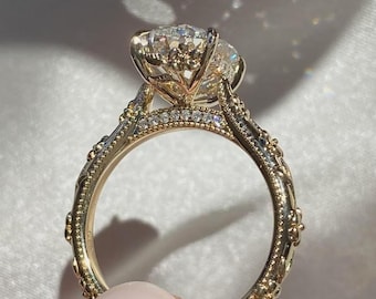Könnte beinhalten: Ein goldener Ring mit einem großen, runden, klaren Mittelstein in einer verzierten Fassung. Die Ringschiene ist mit filigranen Metallarbeiten, Milgrain-Kanten und kleinen, funkelnden Akzentsteinen versehen.