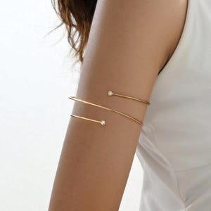 May include: Gold-toned arm cuff with two delicate bands. Each band features a small, sparkling clear gemstone. The jewelry is worn on the upper arm, creating a minimalist and elegant look. The arm cuff is a stylish accessory.
