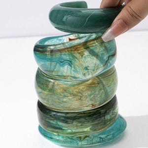 Puede incluir: Una pila de cinco brazaletes de resina veteada en tonos verdes, turquesa y marrón. El brazalete superior se levanta, revelando el diseño de brazalete abierto. Los brazaletes tienen un acabado brillante y un patrón arremolinado.