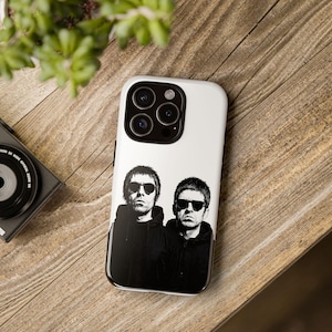 May include: White iPhone case featuring a black and white image of two men wearing sunglasses. The case has a glossy finish and a black border. The phone case is on a wooden surface.
