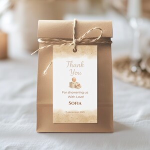 May include: A brown paper gift bag tied with twine, featuring a rectangular tag. The tag reads "Thank You" with a baby graphic and the text "For showering us With Love!" and "SOFIA" and the date "15 december 2025".