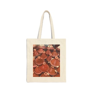 May include: A beige tote bag displaying a print of multiple terracotta tagines. The tagines are in various sizes and shapes, with a deep reddish-brown hue. The bag has a simple design with a rectangular print.