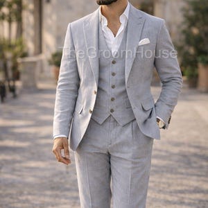 Op de afbeelding: Een lichtgrijs driedelig pak, bestaande uit een jasje, vest en broek, gedragen met een wit overhemd en pochet. Het logo van The Groom House is zichtbaar. Het pak is gemaakt van een lichte stof.