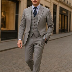 Puede incluir: Un traje de tres piezas gris con rayas finas, que incluye chaqueta, chaleco y pantalón. El traje se lleva con una camisa a rayas azul claro y una corbata azul marino. El hombre camina por una calle adoquinada.