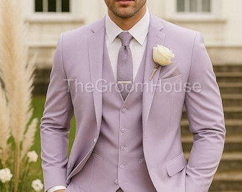 May include: A three-piece lavender suit with a white dress shirt, lavender tie, and a white rose boutonniere. The suit includes a jacket, vest, and trousers. The Groom House is printed on the suit. A silver watch is visible on the wrist.