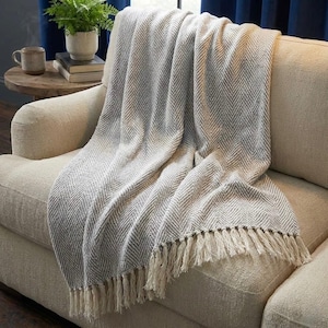 Puede incluir: Una manta de color gris claro y blanco con un patrón de espiga y flecos, colocada sobre un sofá beige. La manta tiene un aspecto suave y texturizado y probablemente está hecha de un material tejido. Los flecos son de color crema.