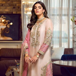 Könnte beinhalten: Ein hellpfirsichfarbenes formelles Kleid mit goldener Stickerei und rosa Akzenten. Das Kleid hat lange Ärmel mit rosa gestreiften Manschetten und aufwendigen goldenen Details. Ein passender rosa Schal liegt über der Schulter. Das Outfit wird mit goldenen Absätzen abgerundet.
